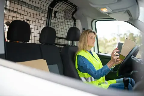 Kobieta w kamizelce odblaskowej sprawdza w telefonie DHL numer przesyłki, trzymając paczkę.