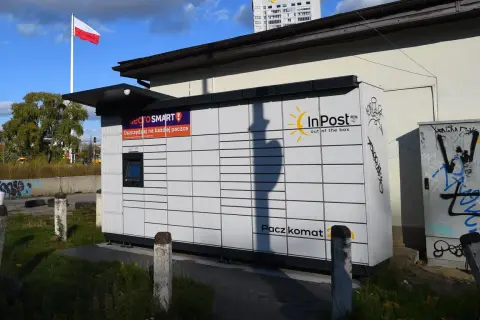 Paczkomat InPost z logo Allegro Smart. W tle polska flaga i budynek. Ile jest paczkomatów w Polsce? Tysiące!