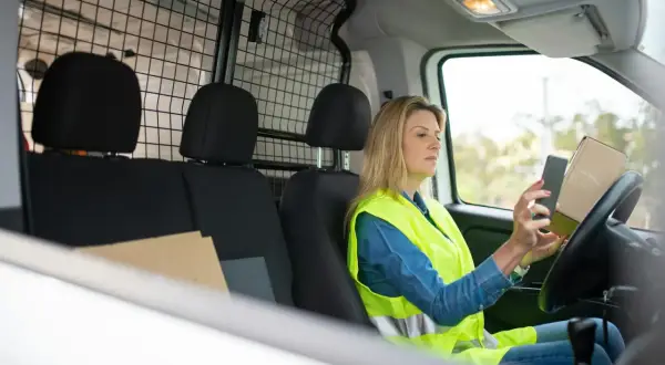 Kobieta w kamizelce odblaskowej sprawdza w telefonie DHL numer przesyłki, trzymając paczkę.