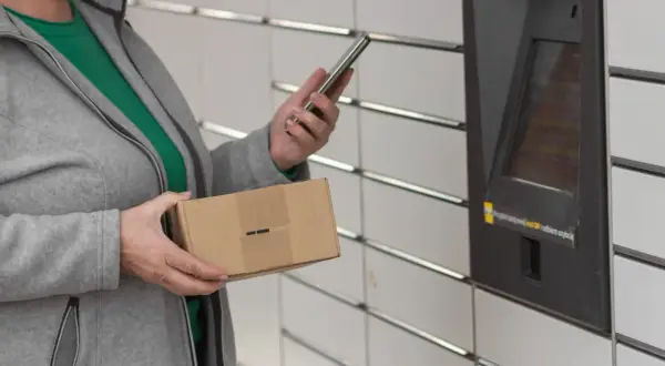 Kobieta z paczką w ręku korzysta z telefonu przy automacie InPost. Szybkie przekierowanie paczki jest teraz łatwiejsze.