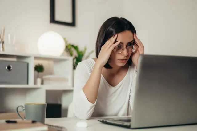 Zestresowana kobieta z okularami masuje skronie, patrząc na ekran laptopa. Brak odpowiedzi na reklamację pogłębia jej frustrację.